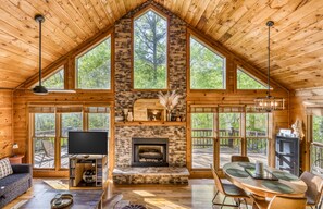 A log cabin with a stone fireplace, wooden floors, a dining area, and large windows offering views of the forest. - Amazing Foliage Views + Mountain View + Hot Tub + Fire Pit + Wrap-around Deck (Cherry Log)