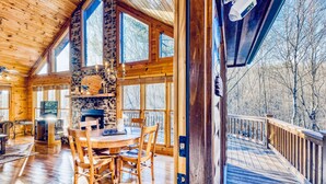 A spacious wooden interior with a large dining table, stone fireplace, and large windows offering views of the surrounding trees.