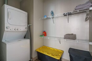 A laundry room with a washing machine, shelves with folded towels, and a trash bin.