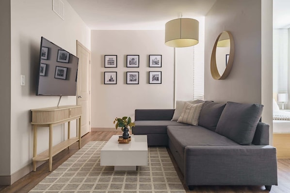 Un salon moderne avec un canapé gris, une table basse blanche et un téléviseur fixé au mur. Des tableaux encadrés ornent le mur, ainsi qu’un miroir rond.