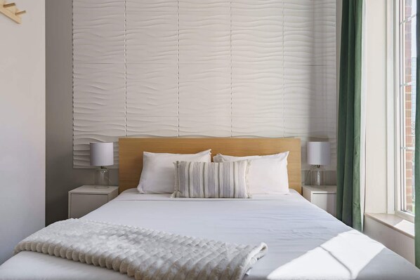 Uma cama bem arrumada com cabeceira de madeira, duas mesas de cabeceira com abajures e uma janela com cortinas.