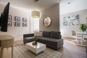 A modern living room with a sofa, coffee table, and a TV. There are framed pictures on the wall, a round mirror, and a plant.