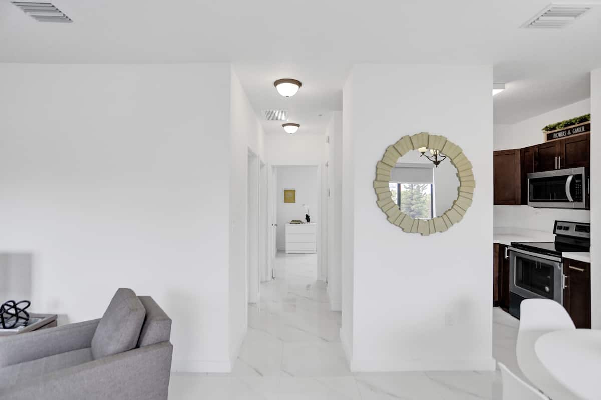 A modern interior with a hallway leading to a kitchen area, featuring white walls, a grey sofa, and a decorative mirror.