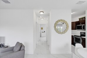 Un intérieur moderne avec un couloir menant à une cuisine, caractérisé par des murs blancs, un canapé gris et un miroir décoratif.