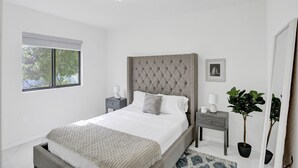A bedroom with a large bed, a nightstand, a dresser, a window with blinds, and a potted plant. -  Renzzi Wynwood Apartments (Miami)