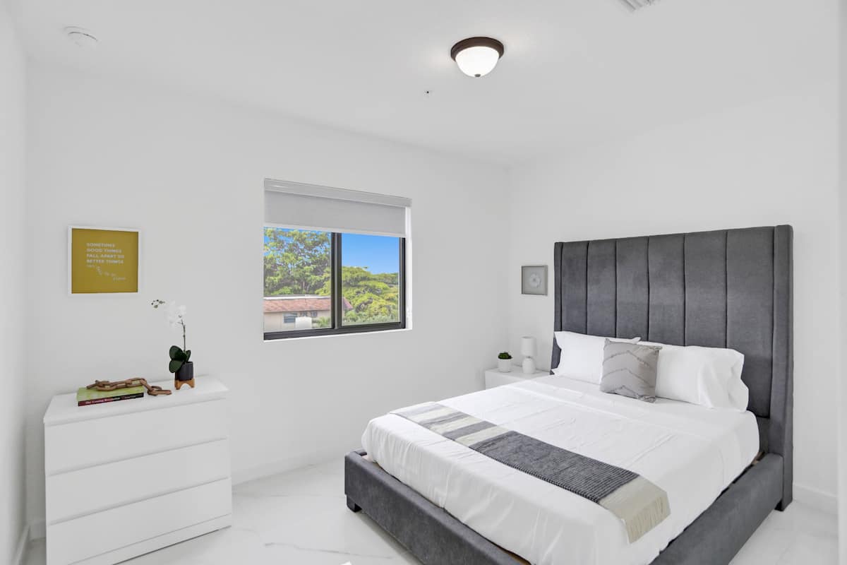 A modern bedroom with a large bed, white nightstands, a window with a view, and a wall-mounted light fixture.