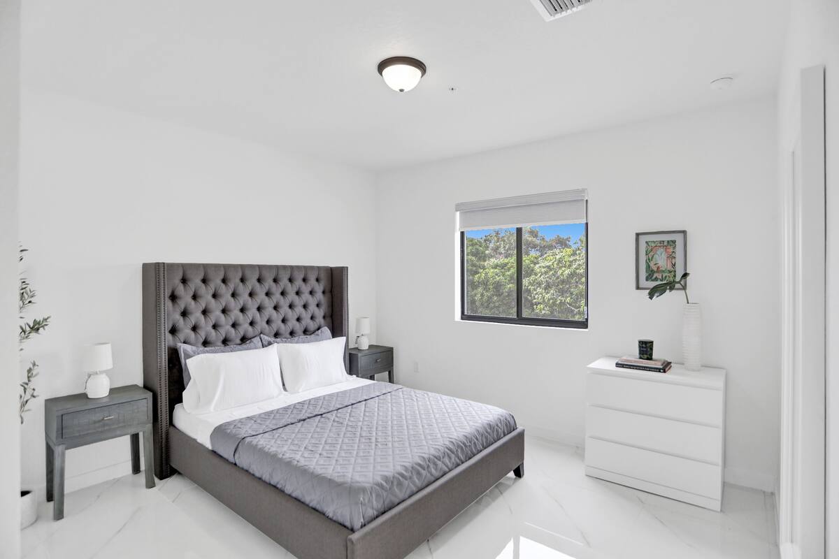 A modern bedroom with a tufted headboard, a large window with a view of trees, a bedside table with a lamp, and a white dresser.