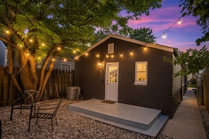 A small, dark-colored building with a white door and windows, surrounded by a wooden fence and trees.