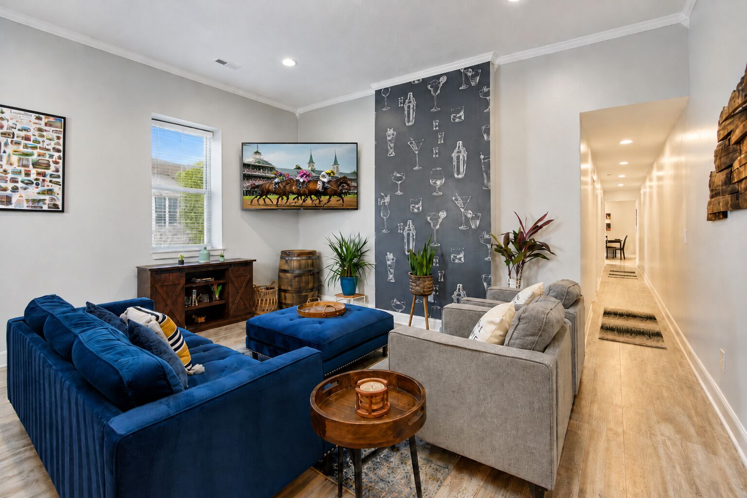 A living room with a blue sofa, a grey sofa, and a wooden coffee table. There is a television mounted on the wall displaying a horse race.