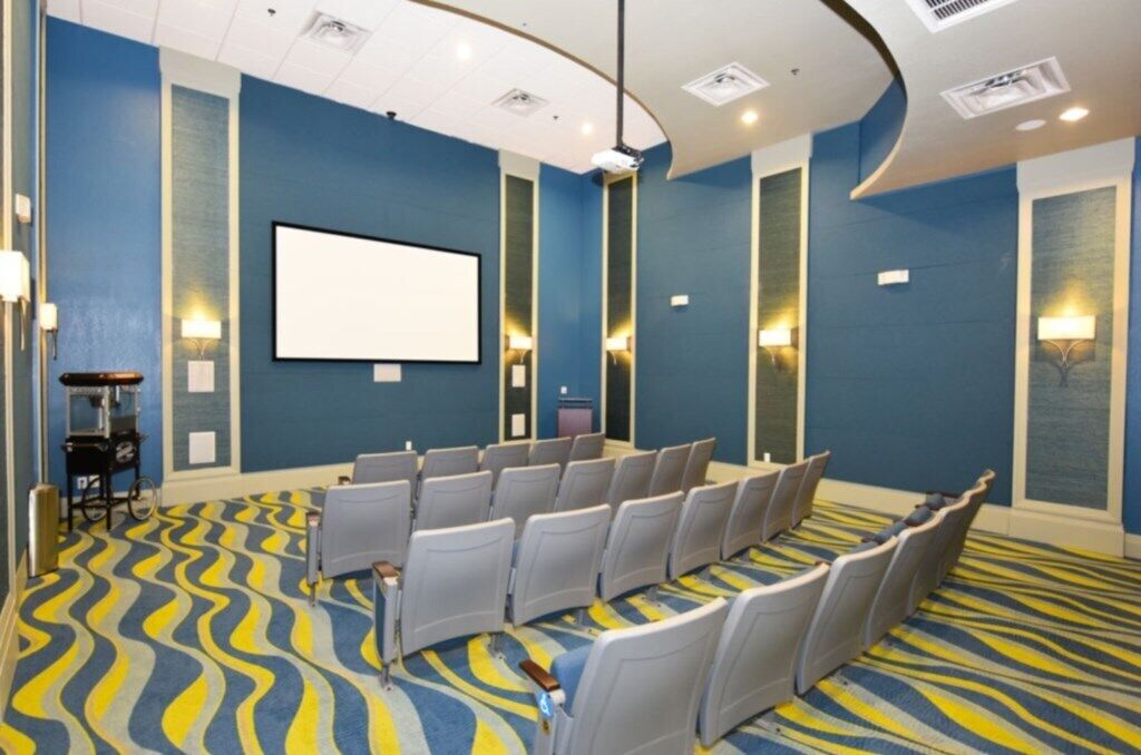 Un salón de conferencias con filas de sillas, una pantalla grande y una alfombra estampada.