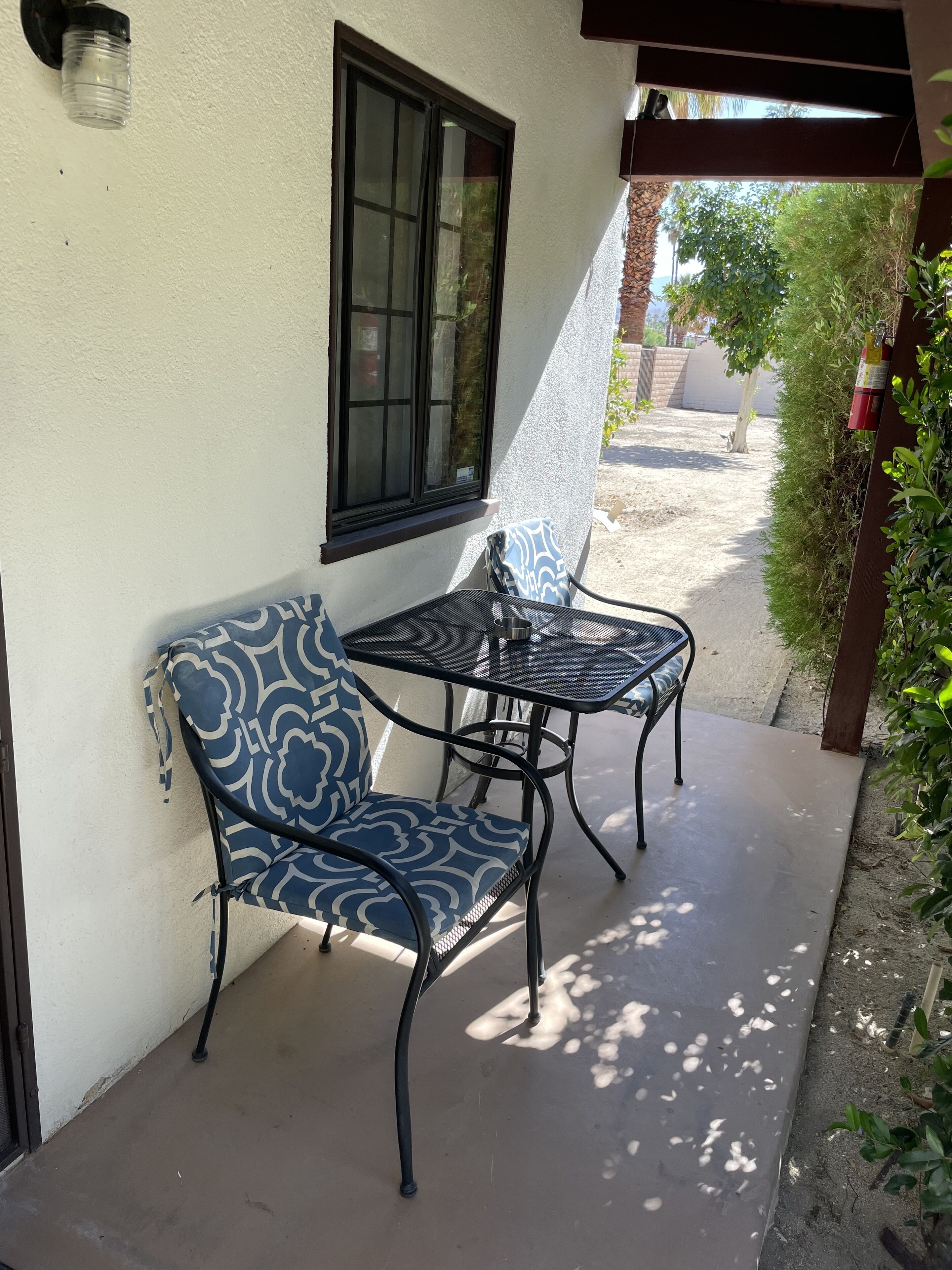 A patio with a blue patterned chair and a metal table, a window, and a fire extinguisher.