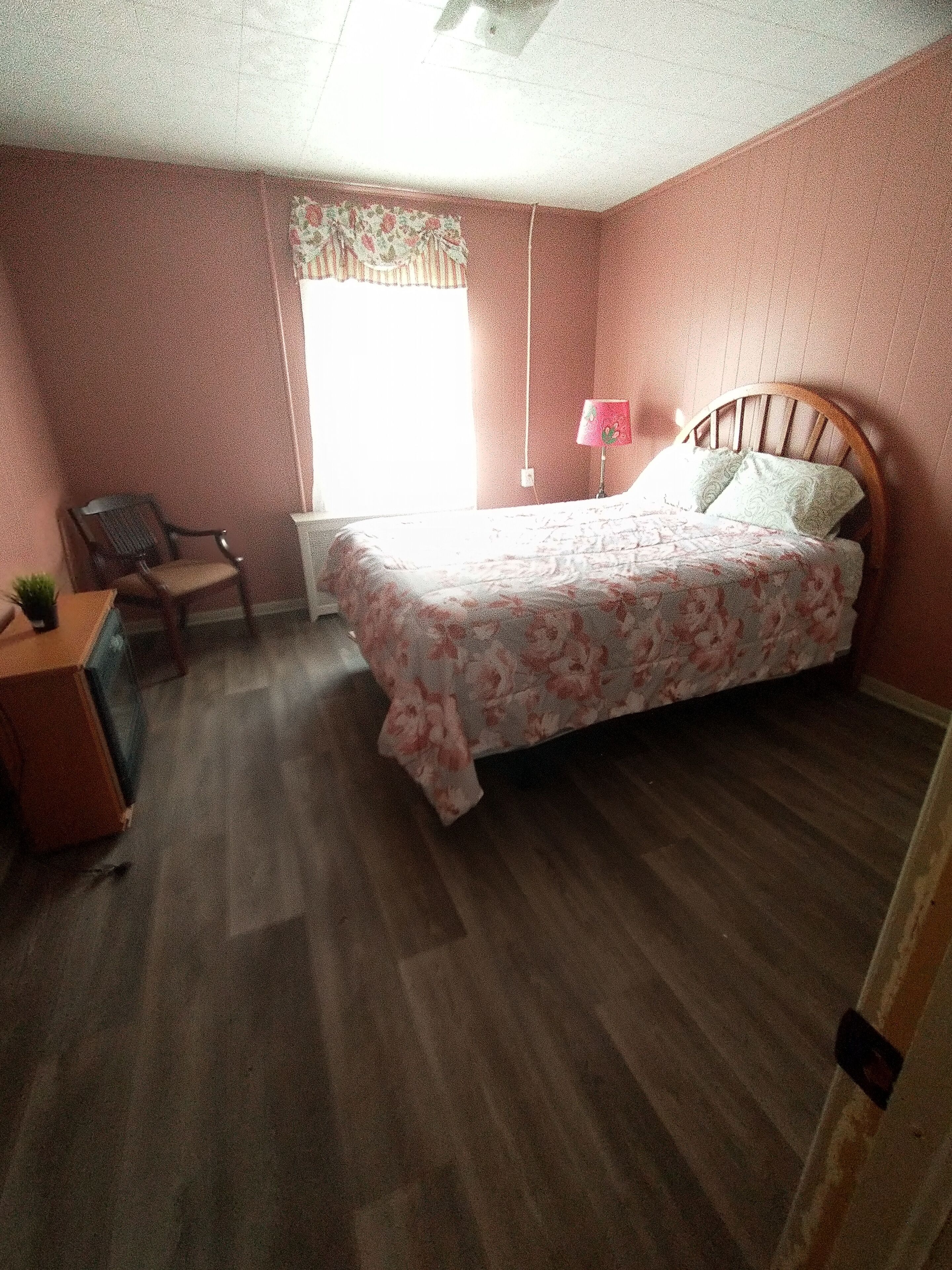 A bedroom with a wooden bed, a desk with a chair, a window with floral curtains, and a small plant on the desk.