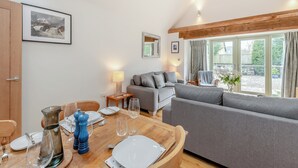 Ein gemütliches Wohnzimmer mit grauem Sofa, einem Holzespeisetisch für zwei Personen und einem großen Fenster mit Vorhängen.