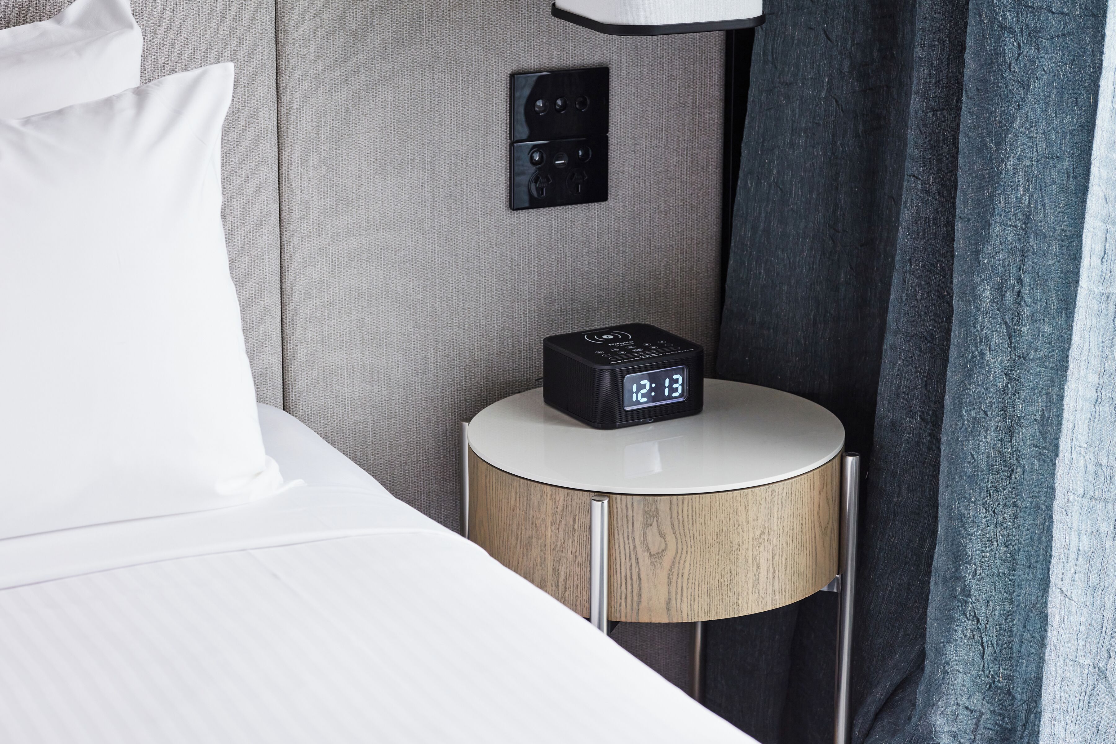 A neatly made bed with white linens, a round bedside table with a wooden finish, a black alarm clock displaying 12:13, and a wall-mounted power outlet.