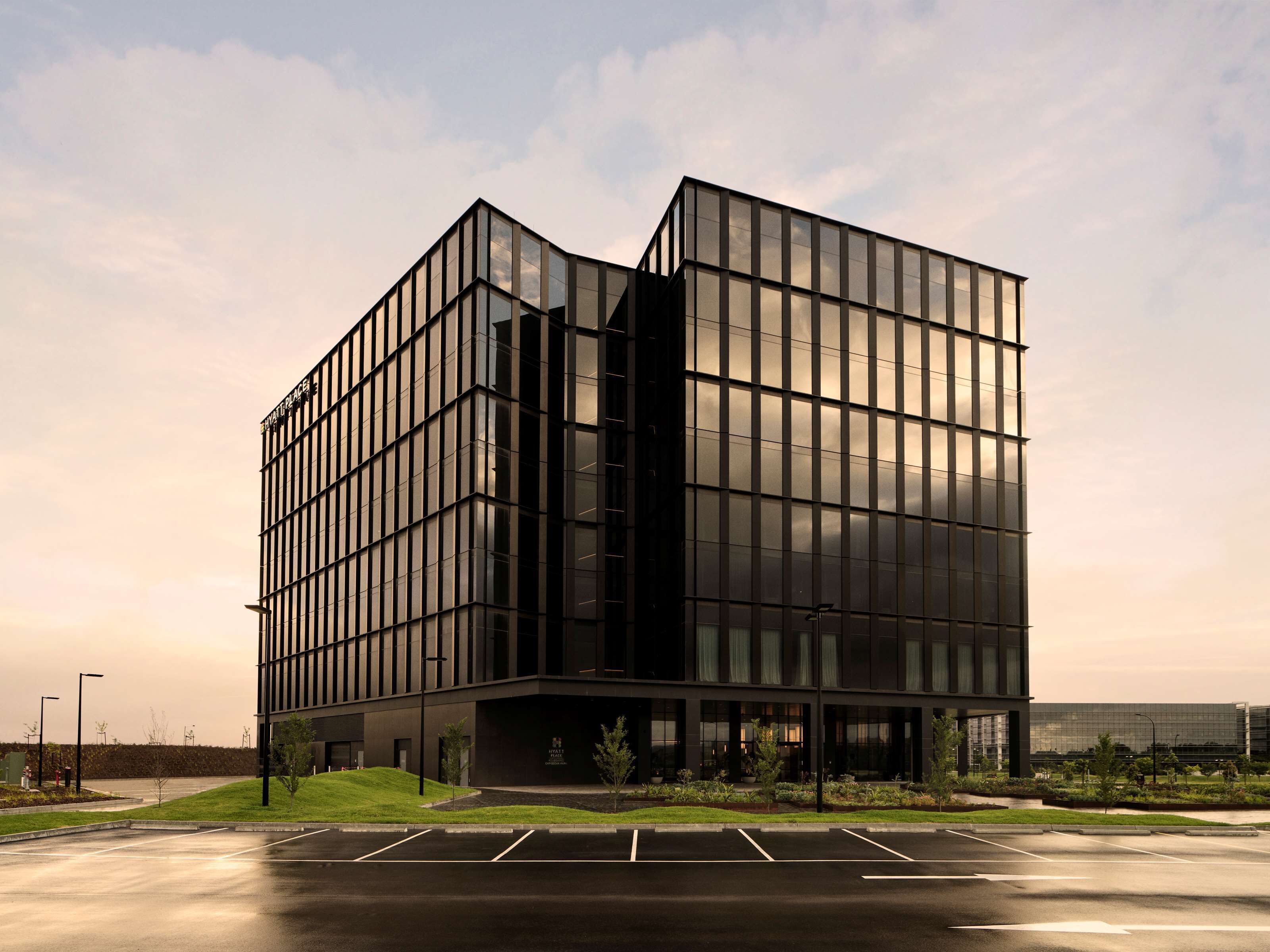 A modern building with a glass facade, a parking lot in front, and a landscaped area with small trees.