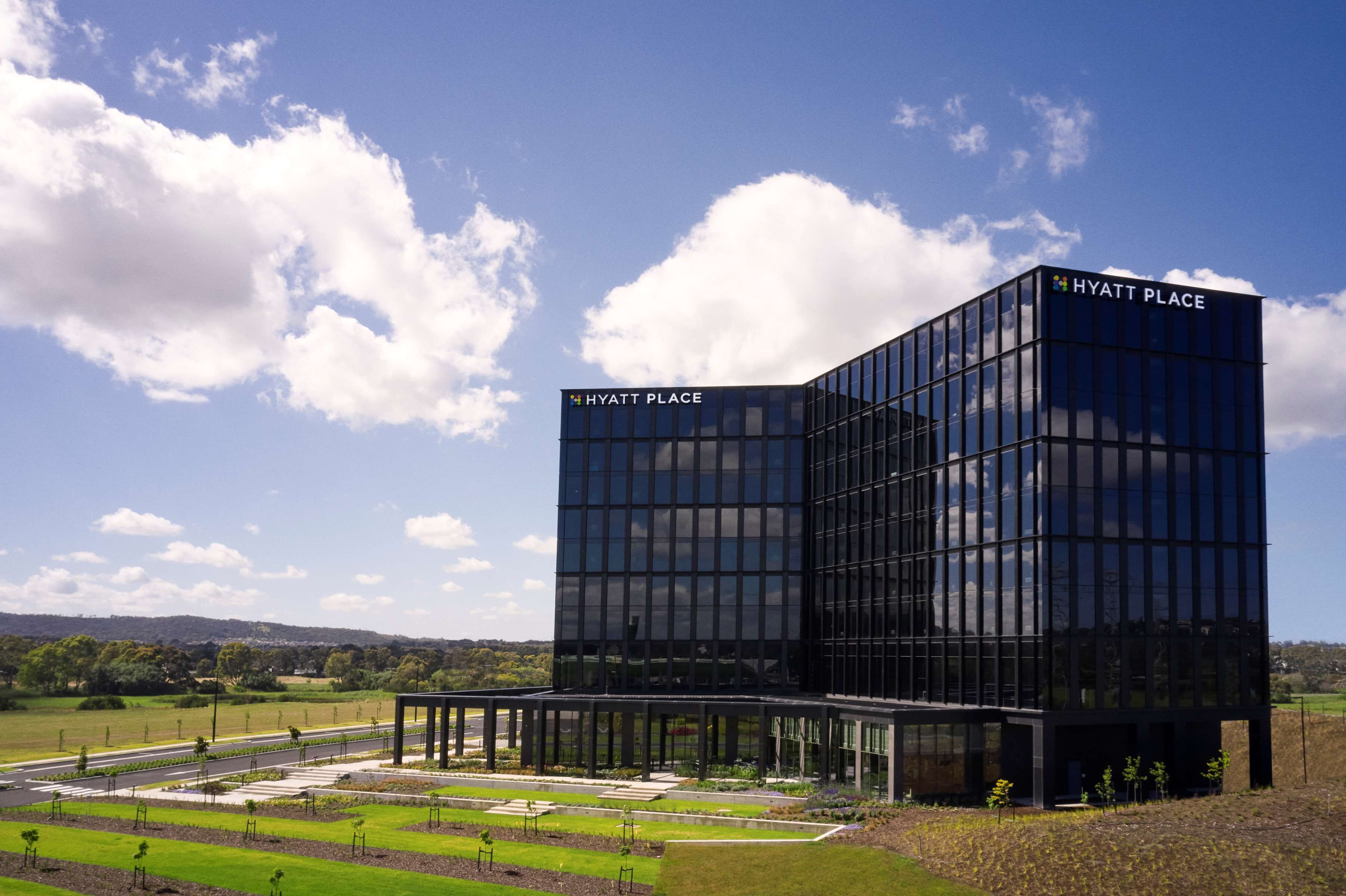 A modern Hyatt Place hotel with a glass facade, surrounded by landscaped gardens and a clear sky.