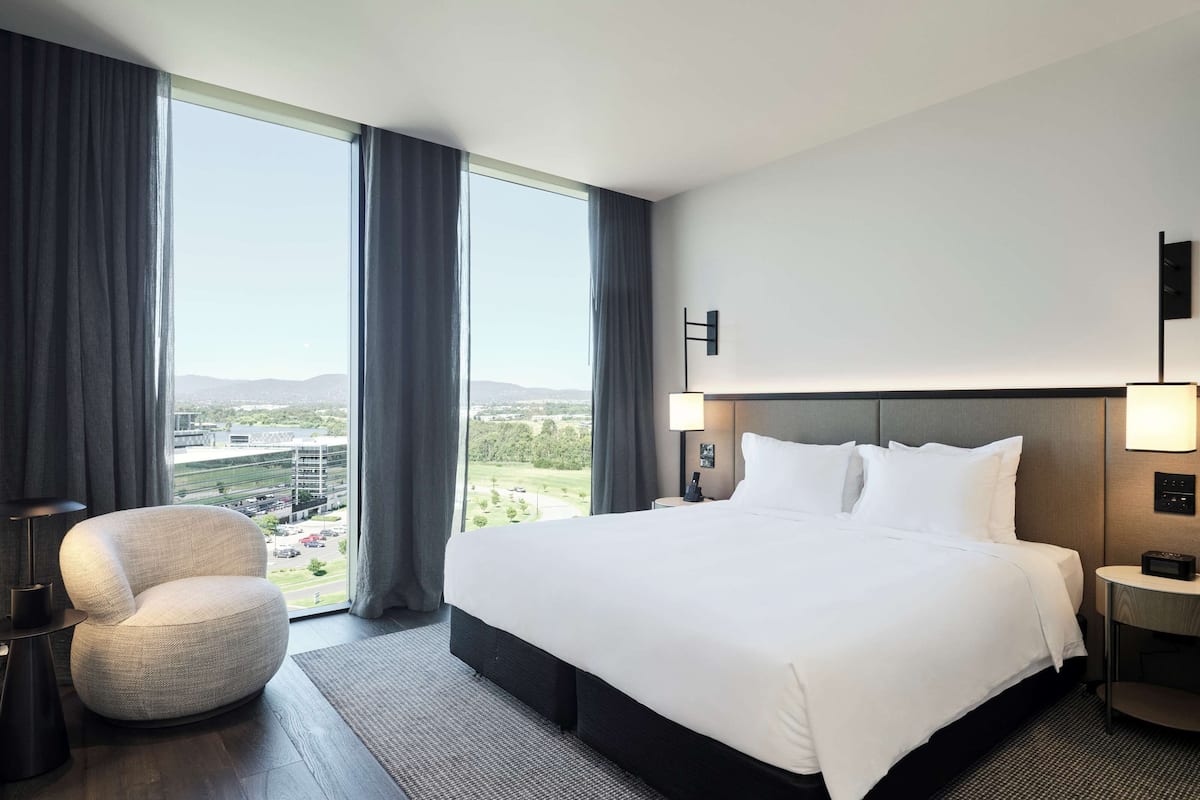 A modern hotel room with a large bed, a round armchair, a nightstand with a lamp, and a view of a cityscape through large windows.