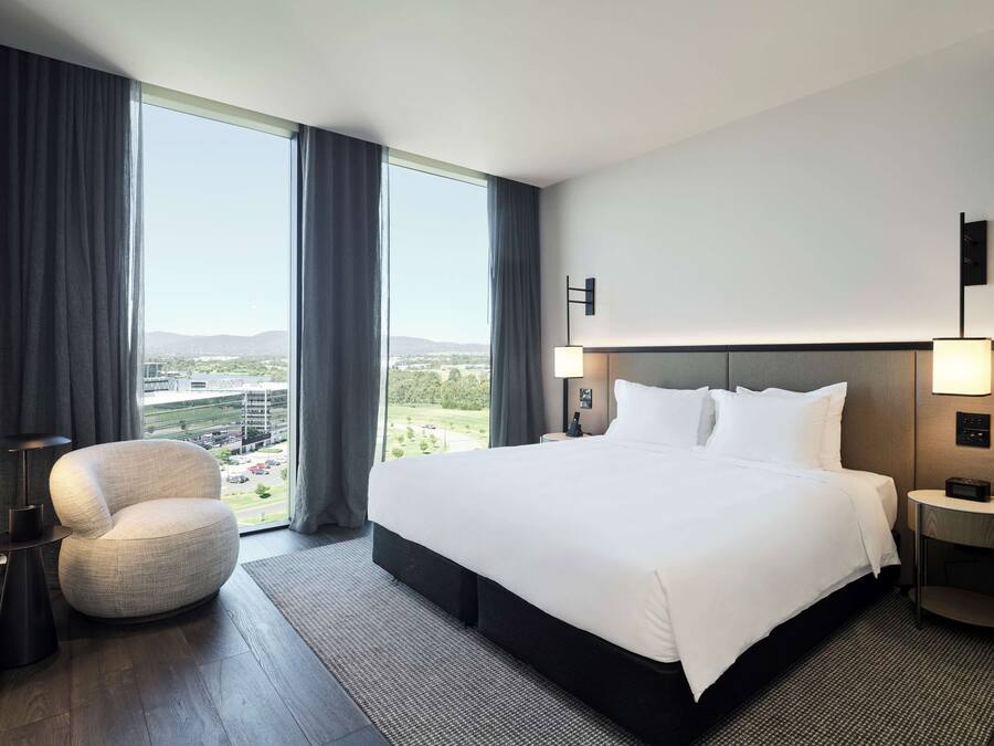 A modern hotel room with a large bed, a round armchair, a nightstand with a lamp, and a view of a cityscape through large windows.