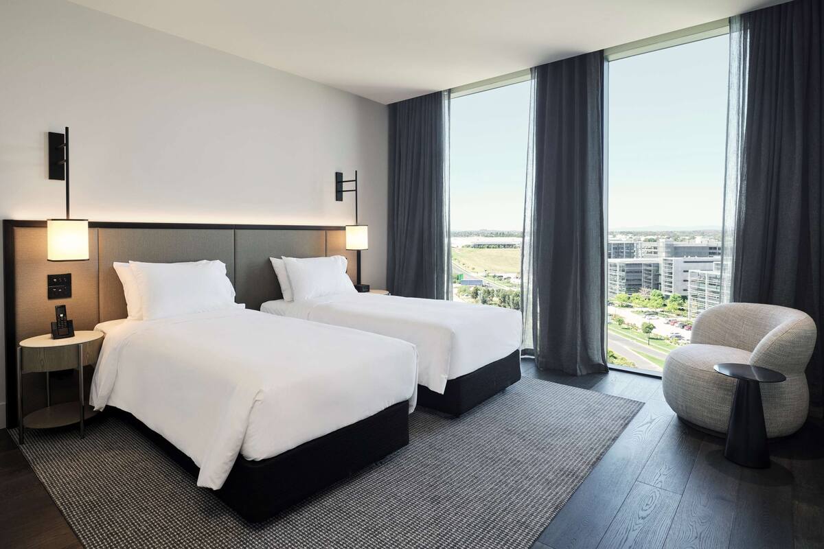 A hotel room with two beds, a chair, a nightstand, and a view of the city.