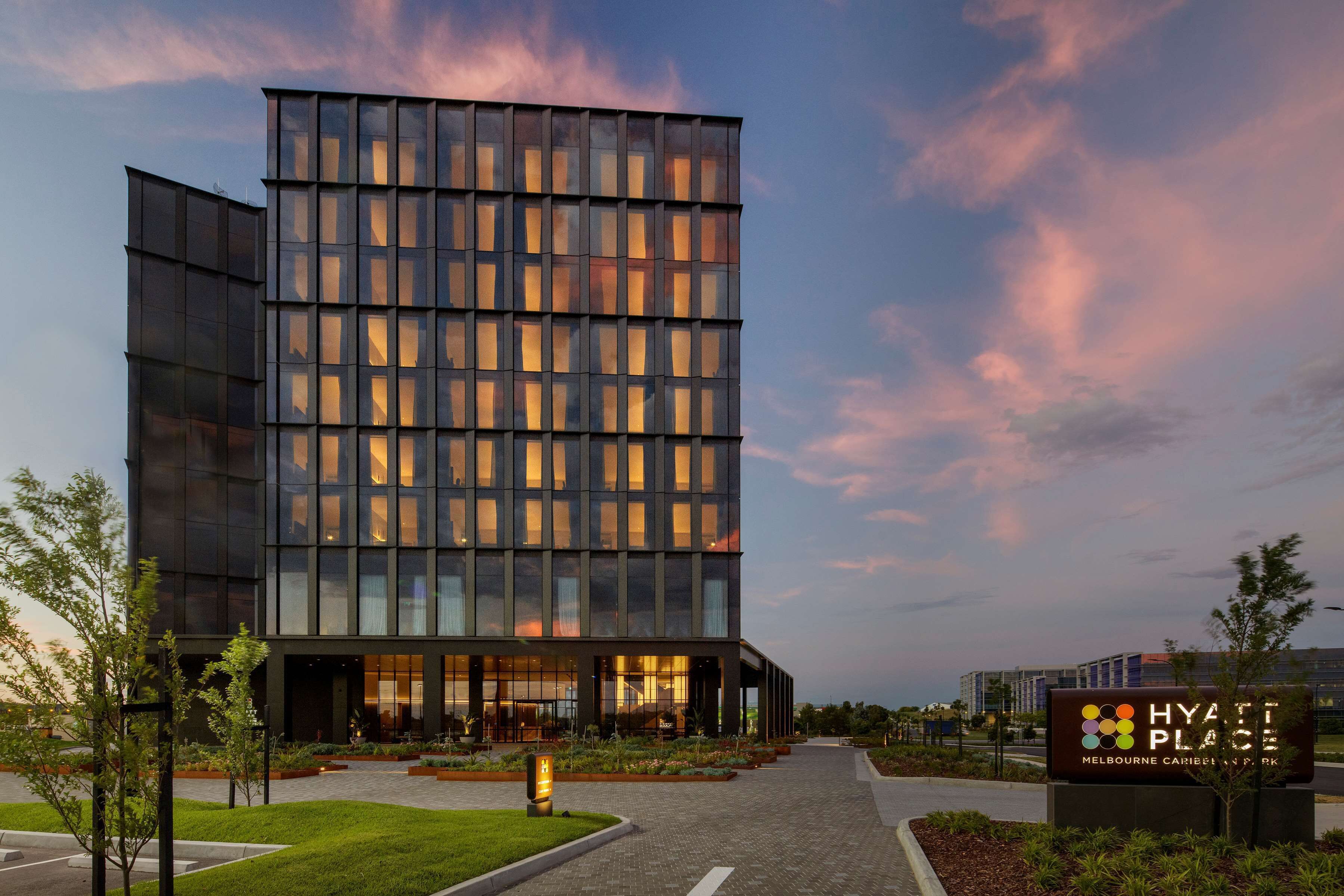 Un moderno edificio de hotel con fachada de vidrio, entrada ajardinada y un letrero que indica que es un Hyatt Place.