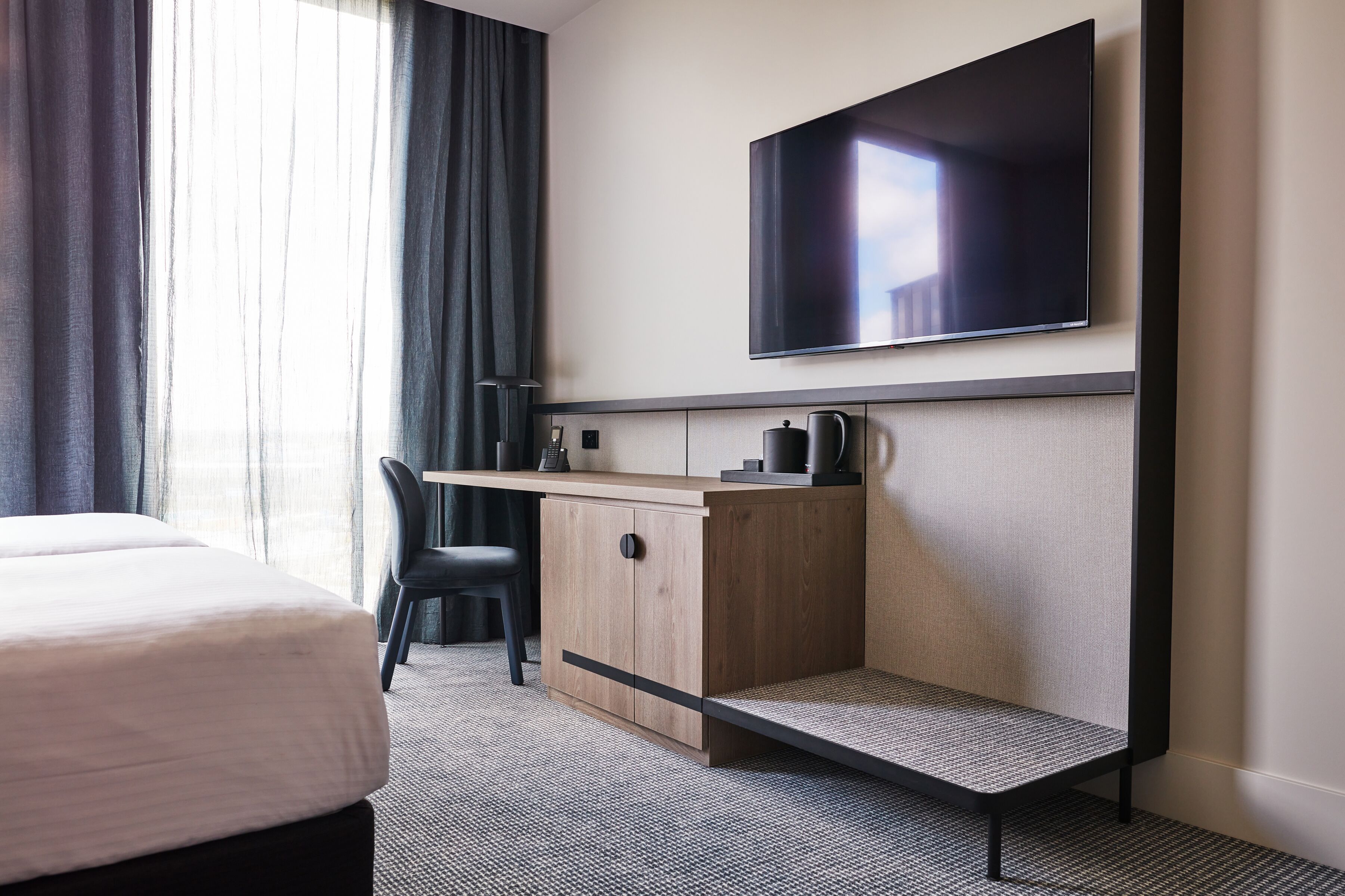 A hotel room with a bed, a desk with a flat-screen TV, a chair, and a small seating area.