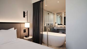 A modern hotel room with a large bed, a bedside table with a lamp, a large mirror, and a freestanding bathtub.