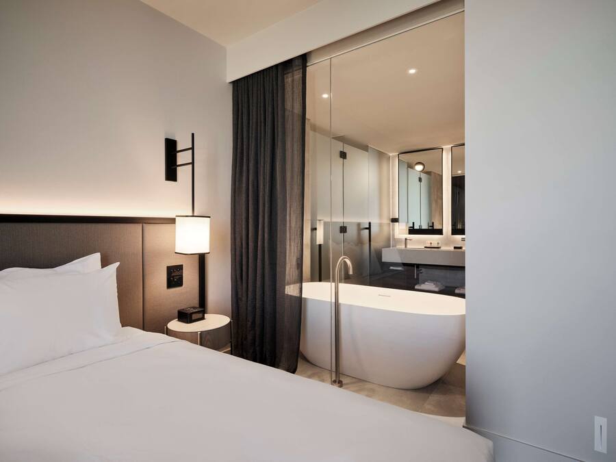 A modern hotel room with a large bed, a bedside table with a lamp, a large mirror, and a freestanding bathtub.