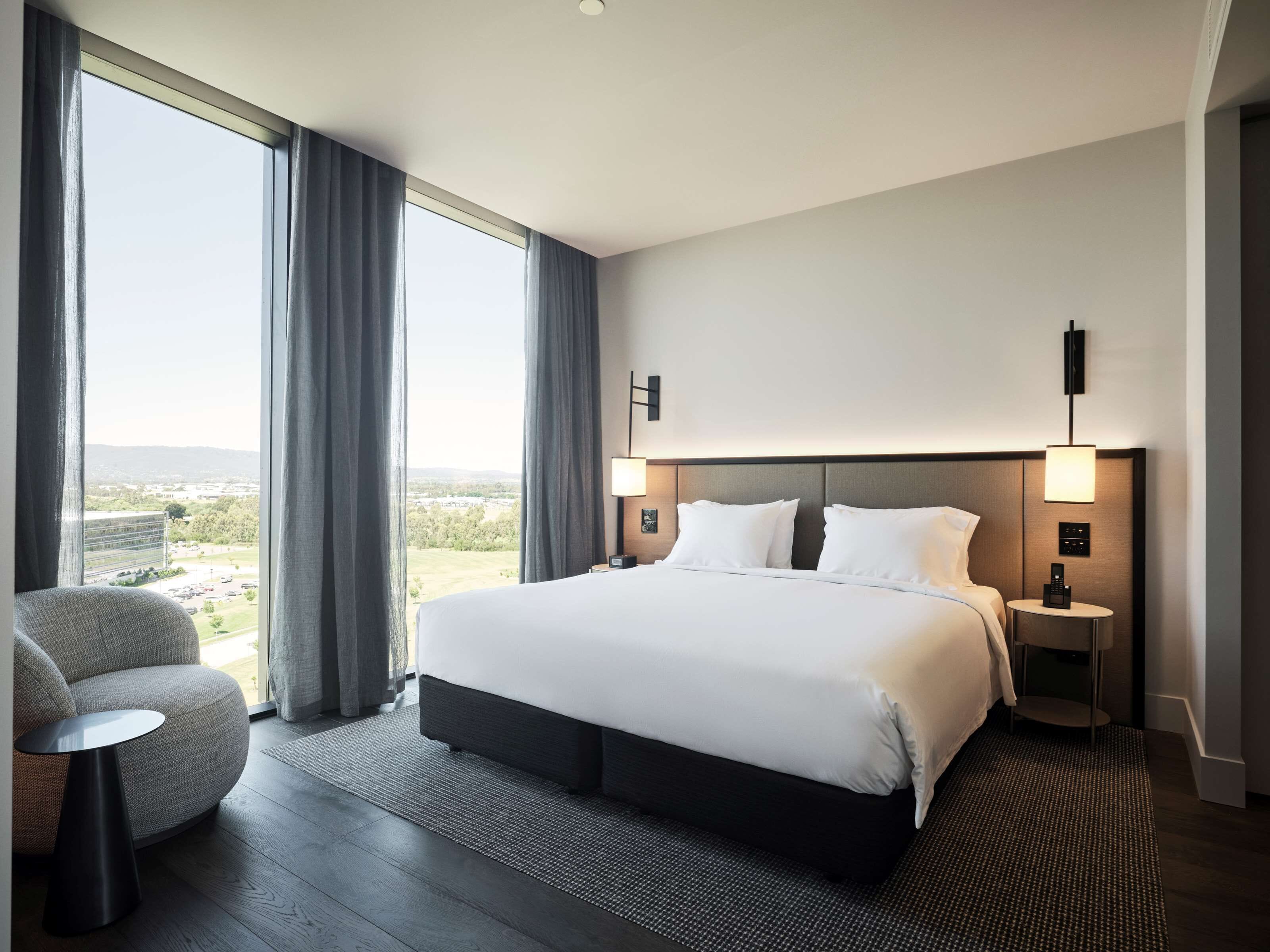 A modern hotel room with a large bed, two bedside tables, a chair, and a view of the cityscape.