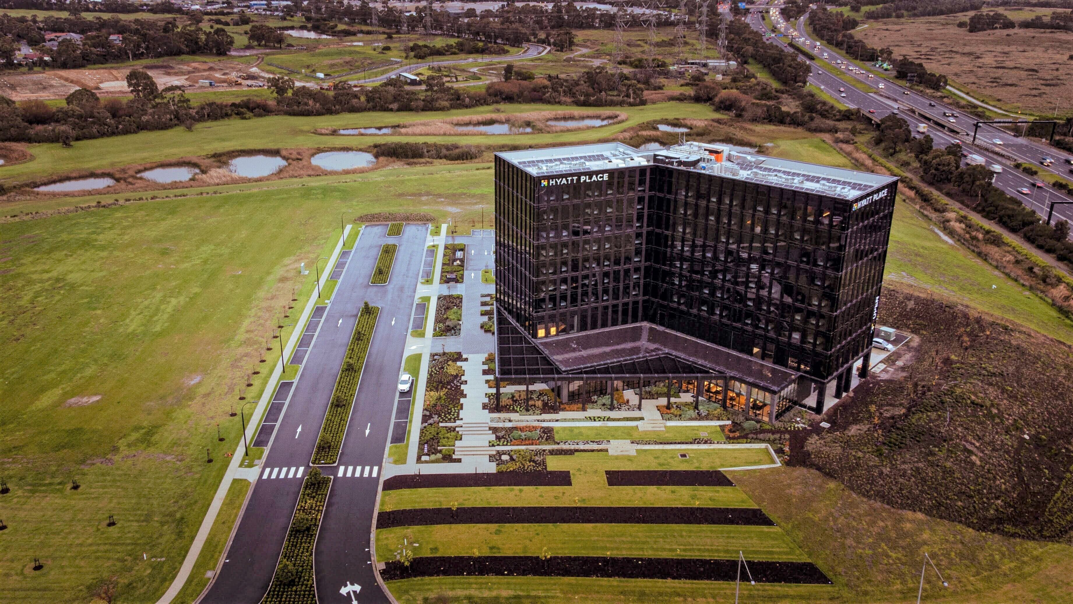 Vista aérea de un moderno hotel Hyatt Place con un amplio estacionamiento, jardines ajardinados y una autopista cercana.