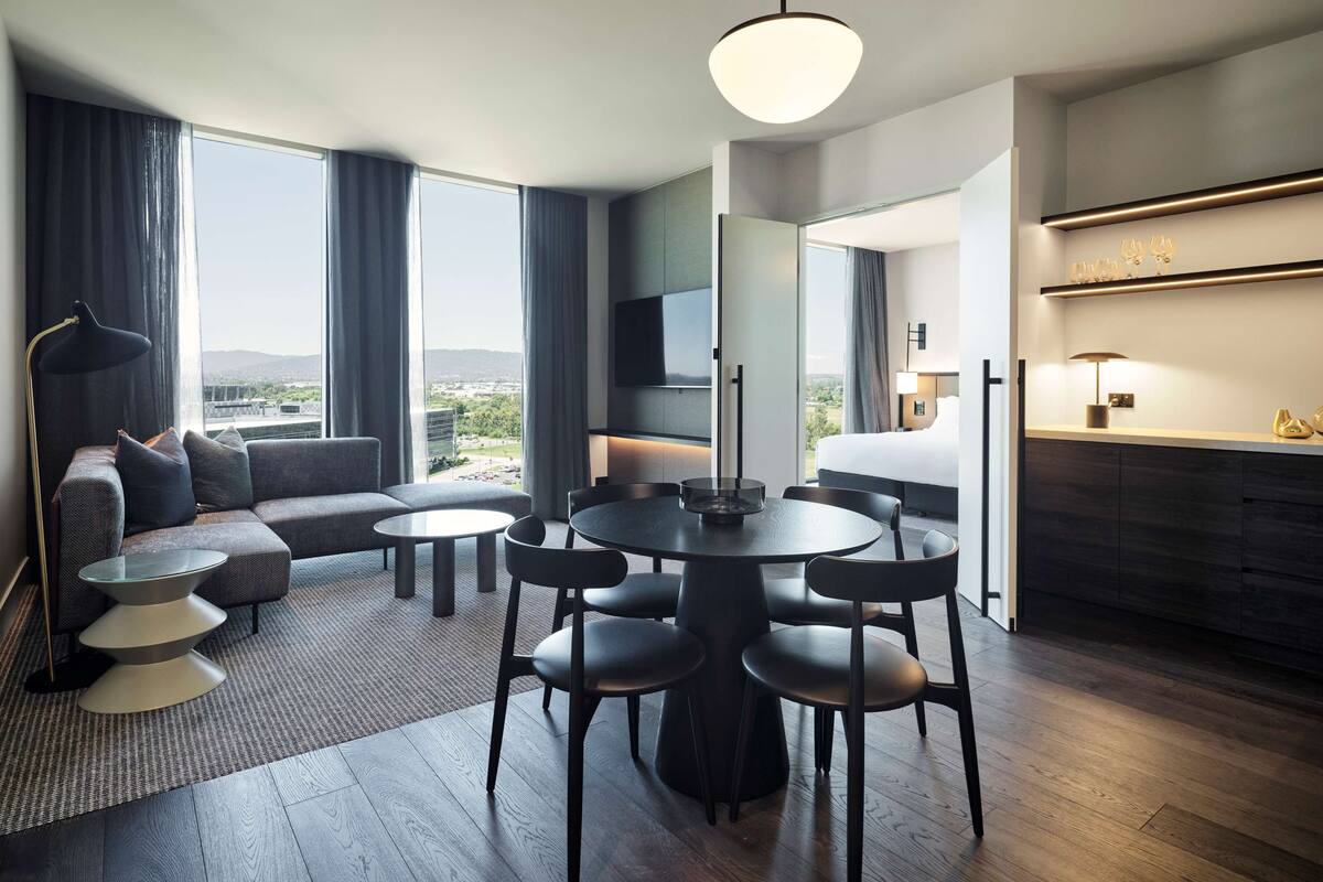 A modern hotel room with a large window, a sofa, a dining table, and a TV.