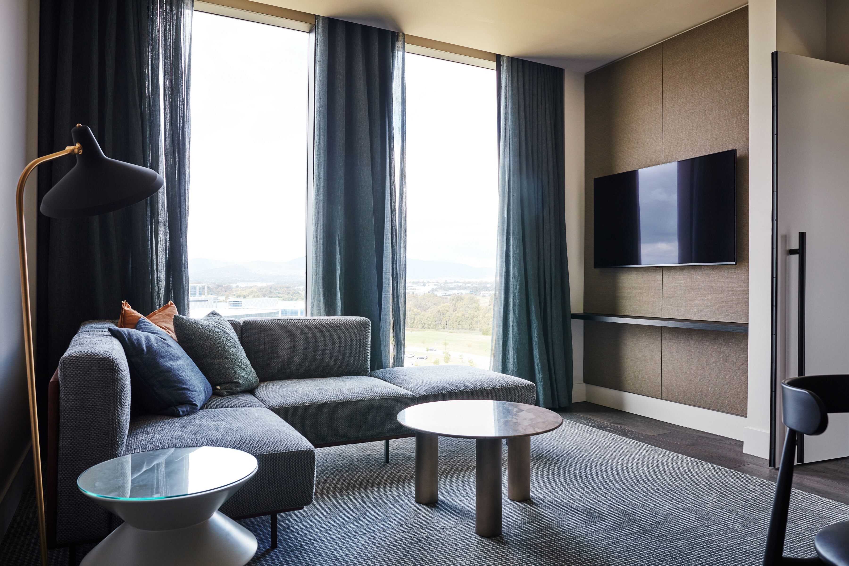 A modern living room with a sectional sofa, two round coffee tables, a floor lamp, and a flat-screen TV mounted on the wall.
