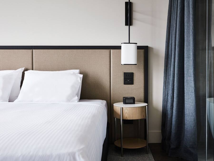 Quarto de hotel com cama, mesa de cabeceira com luminária, painel de controle e área de banho.