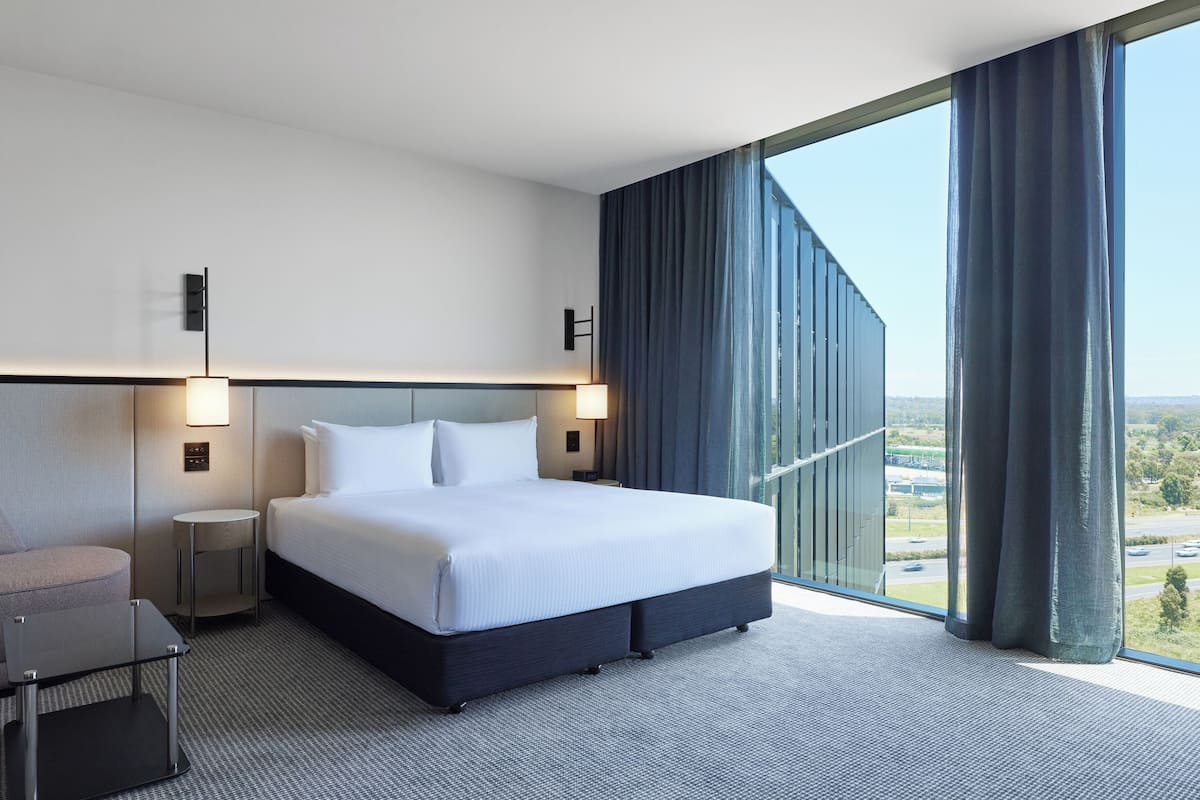 A modern hotel room with a large bed, bedside lamps, a small table, and a view of the city through a large window.