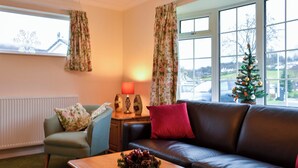A cozy living room with a brown leather sofa, a green armchair, and a wooden coffee table. A small Christmas tree is visible through the window. - Ideally positioned in a peaceful location enjoying extensive fell views. (Keswick)