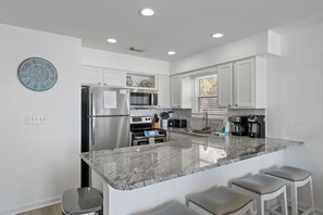 Una cocina moderna con gabinetes blancos, un refrigerador de acero inoxidable, un microondas, una estufa y una encimera de granito. Hay tres taburetes de bar y un reloj en la pared.