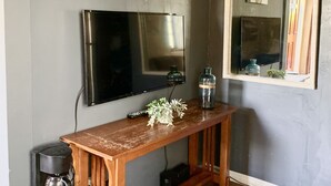 A room with a wooden console table, a flat-screen TV mounted on the wall, a small coffee table, a mirror, and a cactus artwork. - Villa E - a cozy studio in the heart ♥️ of downtown Palm Springs! (Palm Springs)