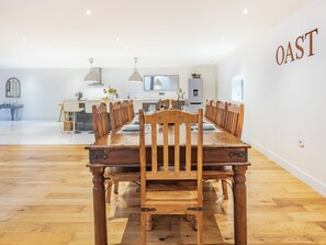A dining area with a long wooden table and chairs, a kitchen in the background, and a wall with the text 'OAST'.