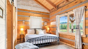 Een houten slaapkamer met een bed, bedlampen, een raam met gordijnen en uitzicht op groen.