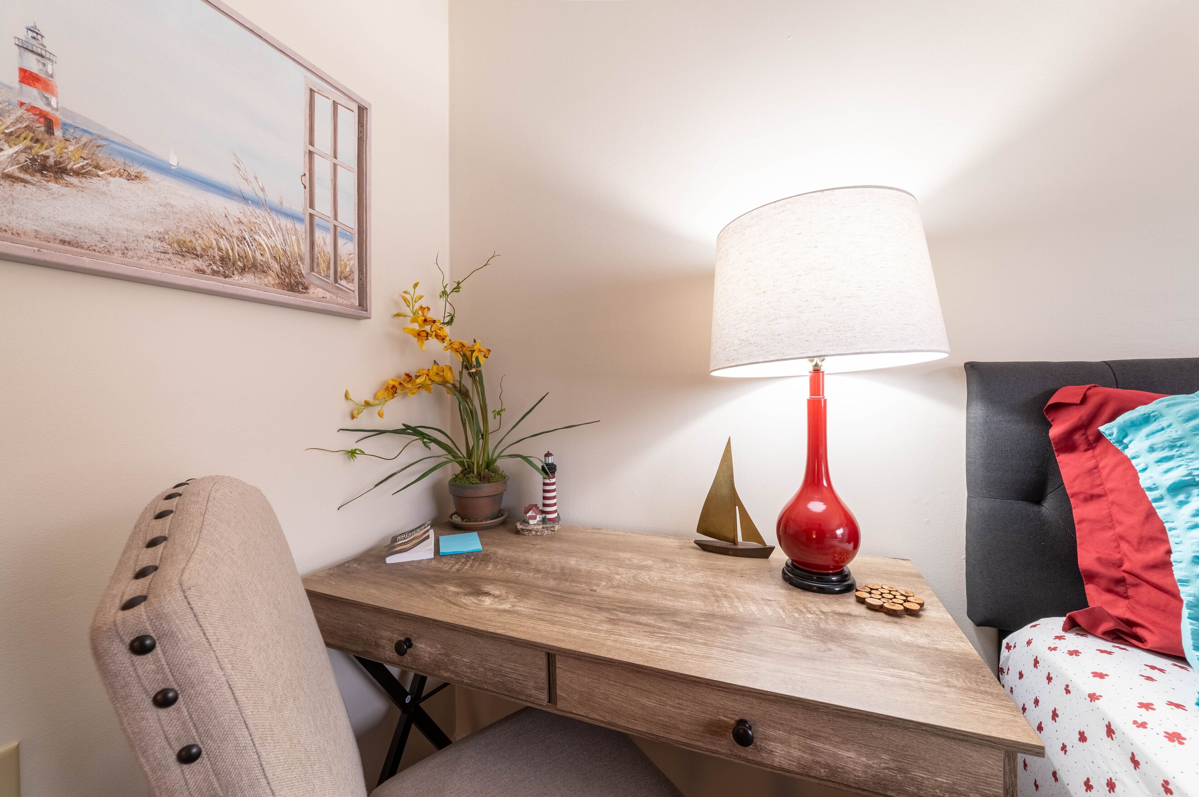 A neatly arranged hotel room with a wooden desk, a lamp, a potted plant, and a bed with a red pillow.