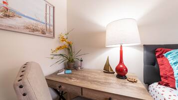 A neatly arranged hotel room with a wooden desk, a lamp, a potted plant, and a bed with a red pillow.