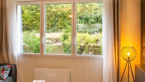 Une pièce avec une grande fenêtre, un radiateur, une petite table sur laquelle se trouve un livre et un lampadaire.