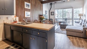 A modern kitchen with dark cabinets and a wooden island, a dining area with chairs, and a living room with a sofa and a lamp. - Walk to Village - Bike Path Access - Pet Friendly 2 BR - Gear Storage - Sleeps 6 (Mammoth Lakes)