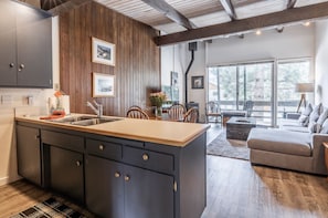 Een moderne keuken met donkere kasten en een houten eiland, een zithoek met stoelen, en een woonkamer met een bank en een lamp.