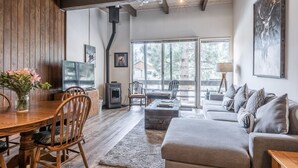 A spacious living room with a wooden dining table, comfortable seating, a fireplace, and large windows offering a view of the outdoors. - Walk to Village - Bike Path Access - Pet Friendly 2 BR - Gear Storage - Sleeps 6 (Mammoth Lakes)