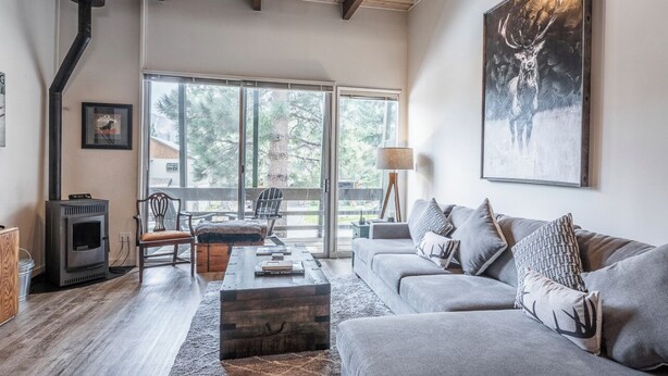 A modern living room with a fireplace, a sofa, a coffee table, and a large window with a view of trees. - Walk to Village - Bike Path Access - Pet Friendly 2 BR - Gear Storage - Sleeps 6 (Mammoth Lakes)