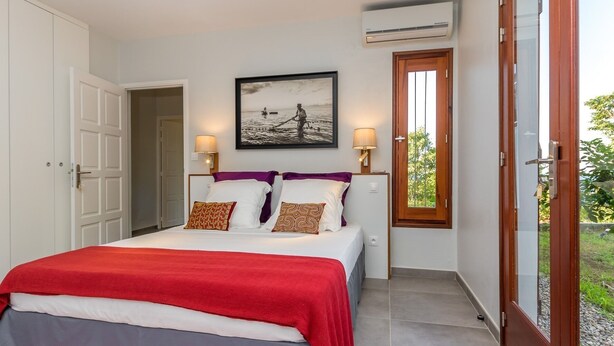 A bedroom with a bed, two bedside lamps, a framed picture, an air conditioning unit, and a door leading to an outdoor area. - Apartment-Royal-Wet room-Partial sea view-Piscine et Salle de Sport (La Trinité)