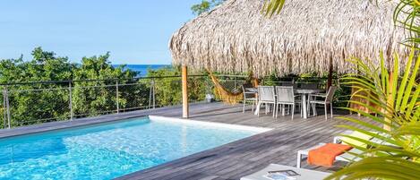 A wooden deck with a pool, a thatched roof structure, and outdoor furniture.