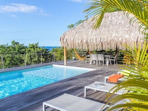 A wooden deck with a pool, a thatched-roof structure, and outdoor furniture.