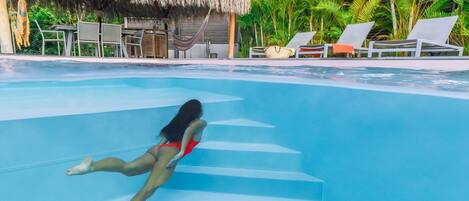 A swimming pool with a person diving, lounge chairs, and a thatched-roof structure.
