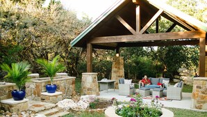 Un área de estar al aire libre con techo, pilares de piedra y mobiliario de mimbre, rodeada por un jardín con diversas plantas y flores.